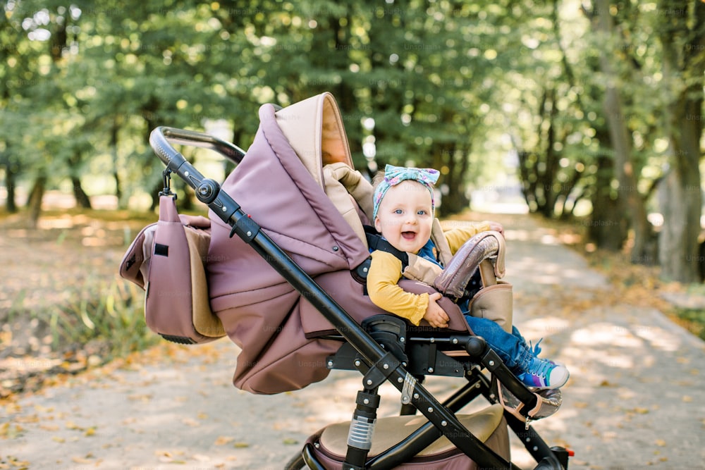 Cute little beautiful baby girl sitting in the pram or stroller and waiting for mom. Happy smiling child with blue eyes