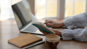 Vista lateral de una chica que escribe en una computadora portátil mientras mira en su cuaderno con una taza de café sobre un fondo de escritorio de madera