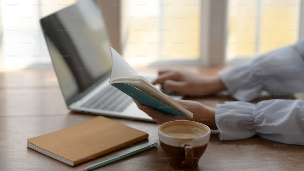 Side view of a girl typing on laptop while looking on her notebook with coffee cup on wooden desk background