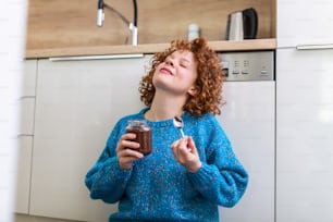 mulher jovem comendo chocolate de um frasco enquanto está sentada no chão da cozinha de madeira. Menina de gengibre bonito entregando-se ao rosto atrevido comendo chocolate espalhado do frasco usando colher saboreando cada bocado