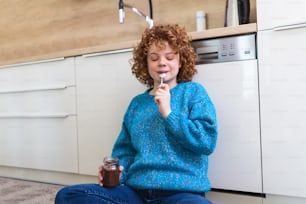 Süßer Ingwer junge Frau in moderner stilvoller Kleidung genießt leckeren Schokoladenaufstrich mit niedlichem Lächeln in der Küche Interier. Junge Frau, die Schokolade aus einem Glas isst, während sie auf dem Küchenboden sitzt.