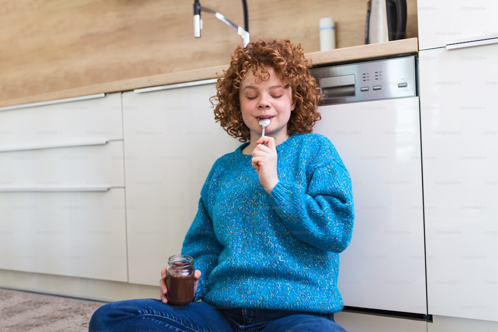 Cute ginger young woman in modern stylish clothes enjoying tasty chocolate spread with cute smile in the cuisine interier. young woman eating chocolate from a jar while sitting on the kitchen floor.