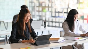 Young beautiful woman with headset working as call center while working/typing on computer tablet and sitting at her working desk surrounded by her colleagues as background.