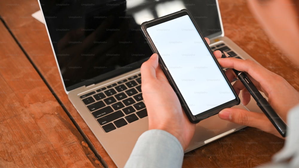 Foto recortada de empresário segurando uma caneta e um smartphone de tela branca na frente do laptop com a mesa de trabalho de madeira como fundo.