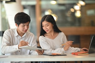 Photo of young creative designer team discussing about work by using tablet and laptop while sitting at the meeting table with modern office as background.