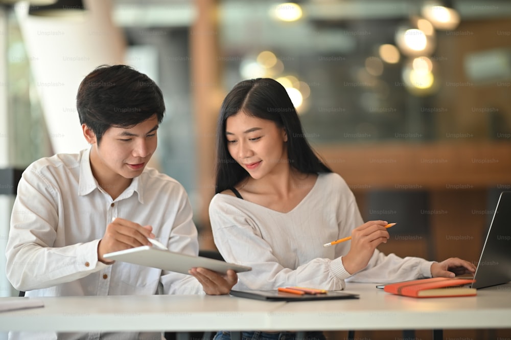 Photo of young creative designer team discussing about work by using tablet and laptop while sitting at the meeting table with modern office as background.