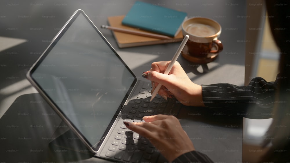 Cropped shot of businesswomen working on blank screen tablet with office supplies and coffee cup in modern dark office room
