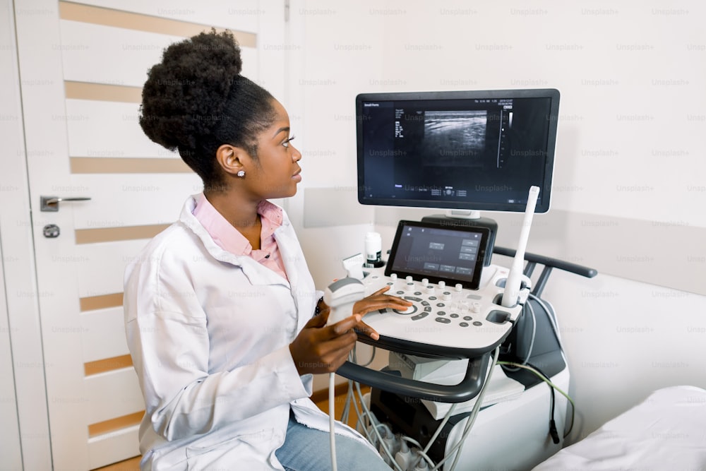 Young african-american female doctor in white coat using ultra ultrasound scanning machine and looking on the screen. African woman doctor working on modern ultrasound equipment