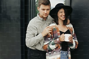 Stylish hipster couple with morning coffee smiling and relaxing on background of modern cabin with big windows in mountains. Happy young family enjoying new home in woods. Space for text