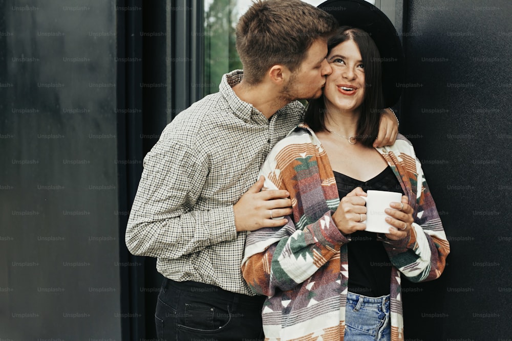 Stylish hipster couple with morning coffee relaxing on background of modern cabin and big windows in mountains. Happy young family of homeowners, enjoying morning in new home in woods