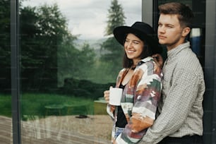 Elegante coppia hipster con caffè mattutino che si rilassa sullo sfondo di una cabina moderna e grandi finestre in montagna. Giovane famiglia felice di proprietari di casa, godendo la mattina nella nuova casa nei boschi