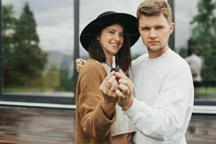 Elegante coppia hipster che tiene insieme la chiave dalla loro nuova casa sullo sfondo della terrazza e delle grandi finestre all'aperto. Giovane famiglia felice in abiti moderni che celebrano l'acquisto della prima proprietà