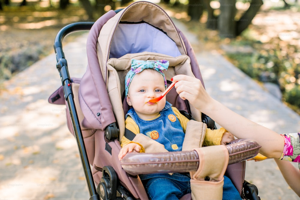 Young attractive mother feeding her cute baby girl, giving her first solid food, healthy vegetable pure from carrot with a plastic spoon sitting on baby stroller carriage and posing smiling.