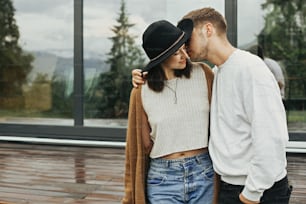 Stilvolles Hipster-Paar, das sich auf einer Holzveranda umarmt und sich in einer modernen Hütte in den Bergen entspannt. Glückliche junge Familie in modernen Outfits, die sich auf dem Hintergrund der Terrasse und des großen Fensters umarmen