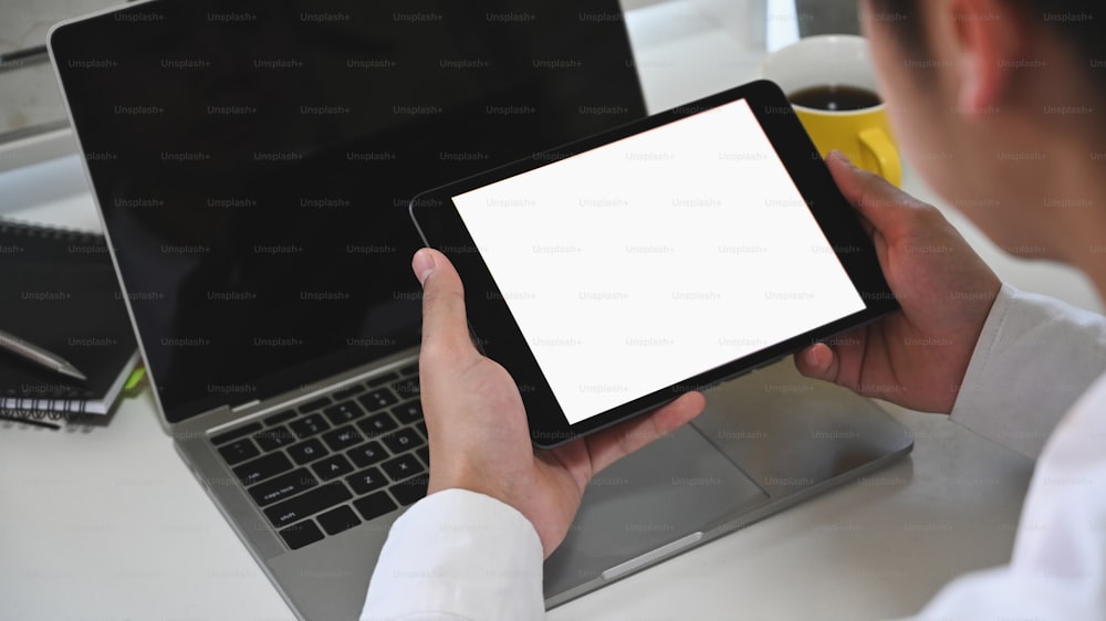Cropped shot business man holding white blank screen tablet at the modern working desk with black blank screen laptop and working desk as background.