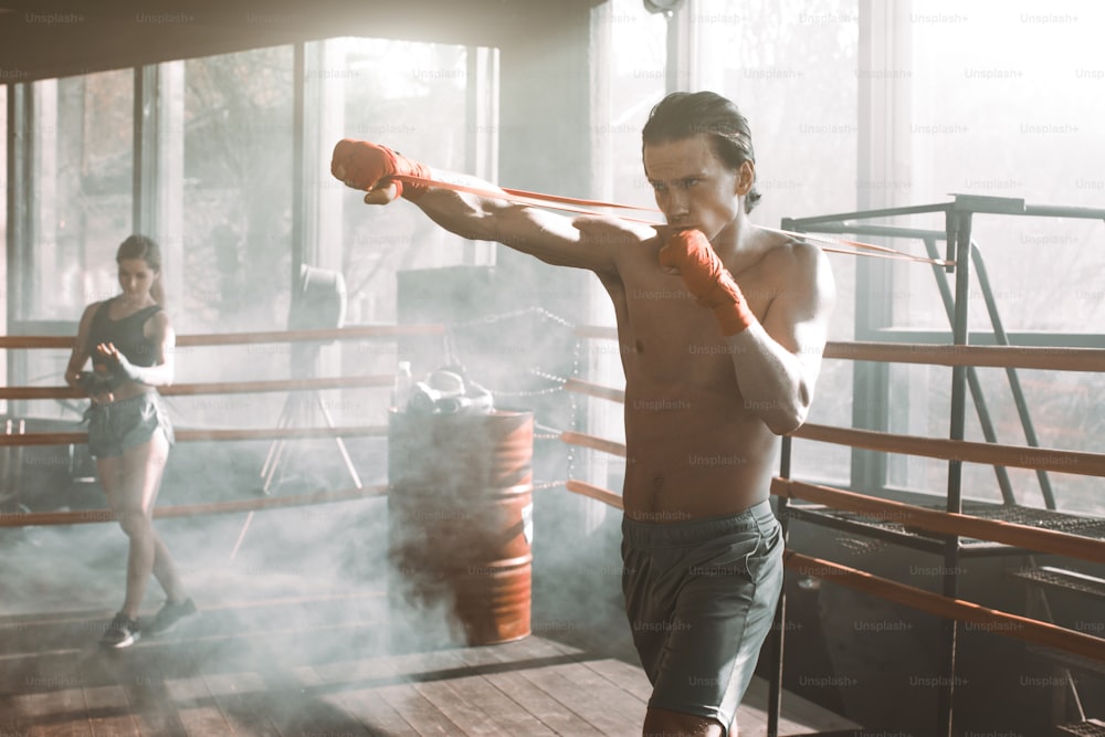 Uomo bello che si allena con la fascia di resistenza sul ring di boxe in palestra. Muscoli delle braccia e della schiena perfettamente modellati.