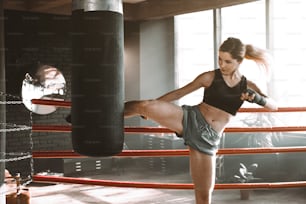 Une jeune femme s’entraîne sur un ring de boxe avec un sac de frappe lourd. Portrait d’un mannequin sexy s’entraînant dans une salle de sport vintage loft. Coups de pied ou coup de genou.