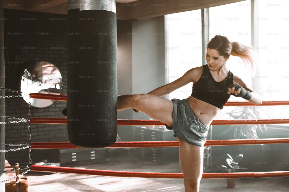 젊은 여자는 무거운 펀칭 가방으로 권투 링에서 훈련합니다. 로프트 빈티지 체육관에서 섹시한 패션 모델 운동의 초상화. 풋 킥 또는 무릎 스트라이크.