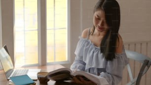Plan recadré d’une fille lisant un livre tout en s’asseyant à côté de la fenêtre dans une pièce confortable avec ordinateur portable et papeterie