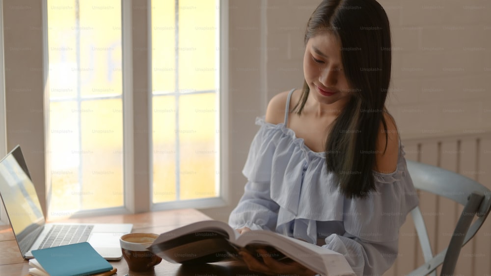 Foto recortada de una niña leyendo un libro mientras está sentada junto a la ventana en una habitación cómoda con computadora portátil y artículos de papelería