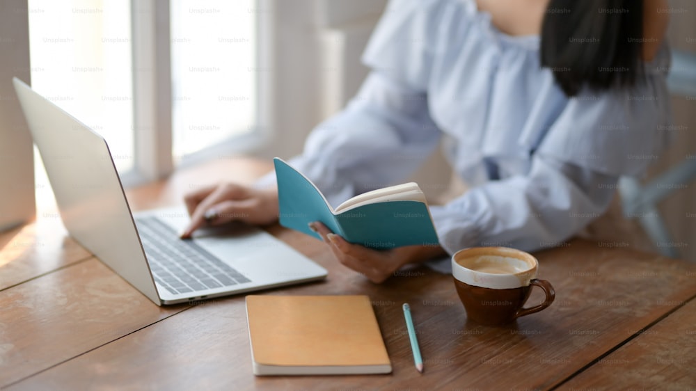 Close up de uma menina digitando no laptop enquanto olha em seu caderno com papelaria e xícara de café no fundo da mesa de madeira