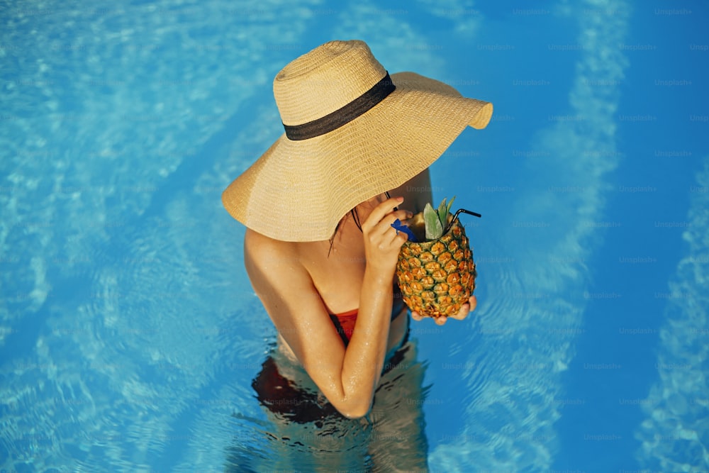 Beautiful young woman in hat drinking delicious cocktail from pineapple and relaxing in pool, summer vacation. Girl enjoying warm sunshine, swimming in pool on rooftop in luxury tropical resort