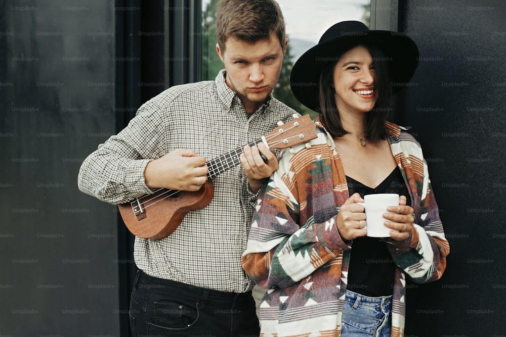 Stylish hipster couple with morning coffee and ukulele smiling, relaxing on background of modern cabin with big windows in mountains. Happy young family enjoying vacation in woods