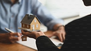 Cropped shot of House broker holding the sample house model in hand with his customer while writing on paper/ signing an agreement at the wooden table. Loan,Debt, selling/buying agreement concept.