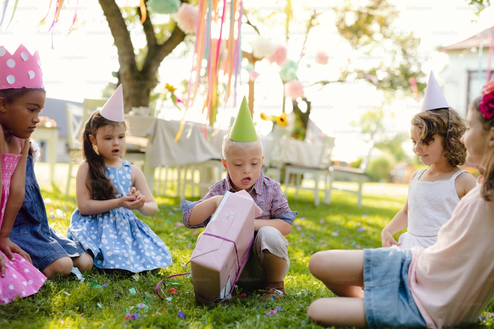 Down-Syndrom-Kind mit Freunden auf Geburtstagsfeier im Freien im Garten, Geschenke öffnen.