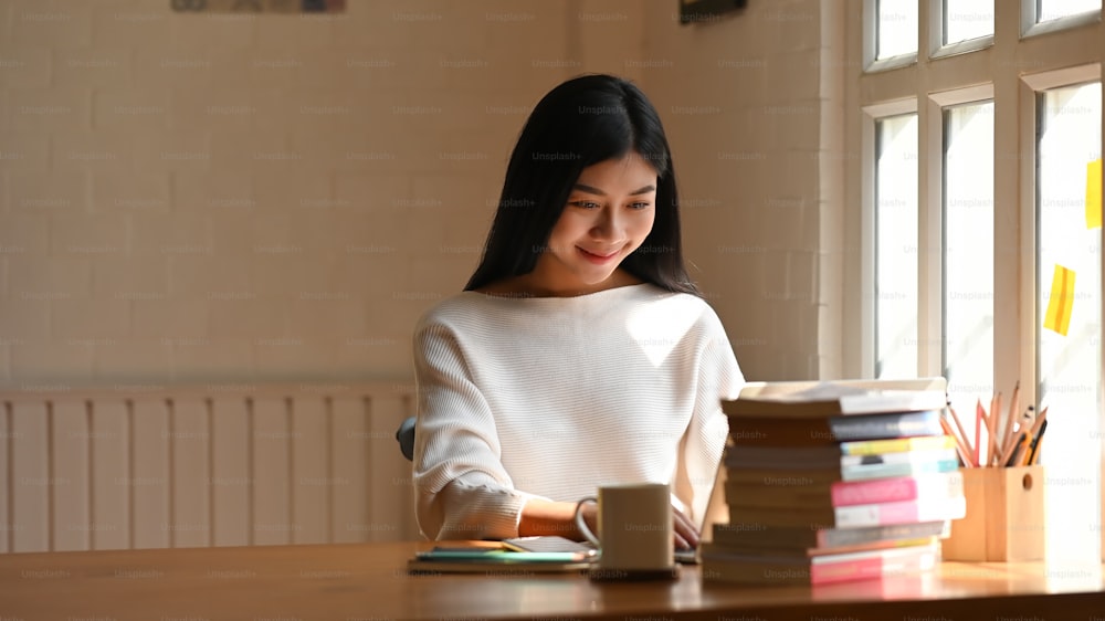 흰색 면 셔츠를 입은 젊은 아름다운 여성이 벽을 배경으로 한 현대적인 나무 테이블에 책, 연필꽂이, 커피 컵 더미 앞에 앉아 노트북 키보드에 타이핑하고 있다.