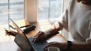 컴퓨터 태블릿을 사용하는 동안 스타일러스 펜을 들고 편안한 거실과 창문을 배경으로 아침에 현대적인 작업 책상에 앉아 있는 자른 샷 여성.
