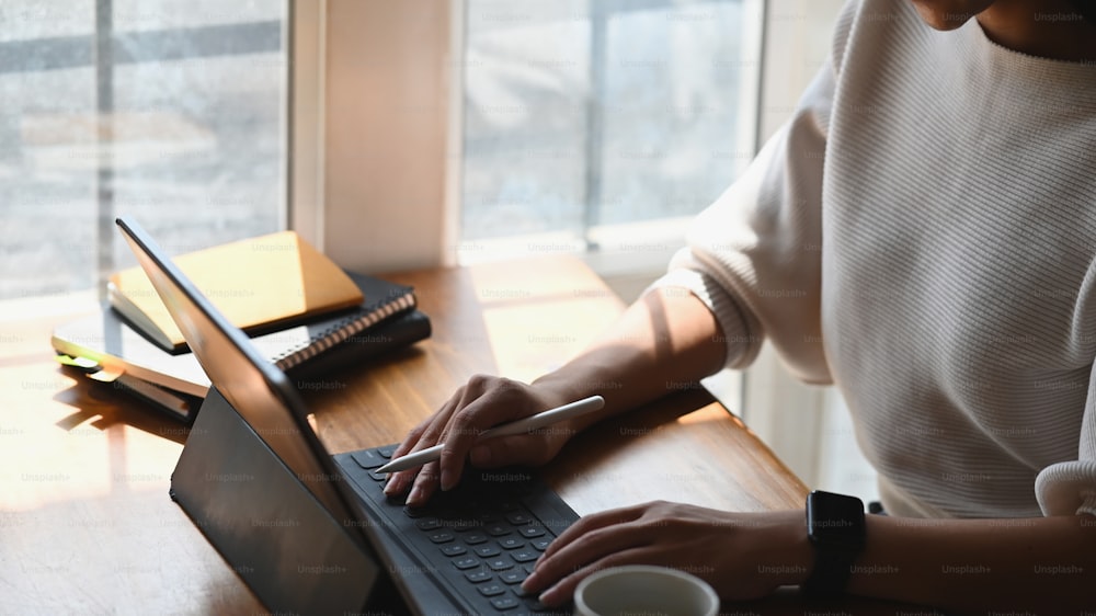 Cropped shot woman holding a stylus pen while using a computer tablet and sitting at the modern working desk in the morning with comfortable living room and windows as background.