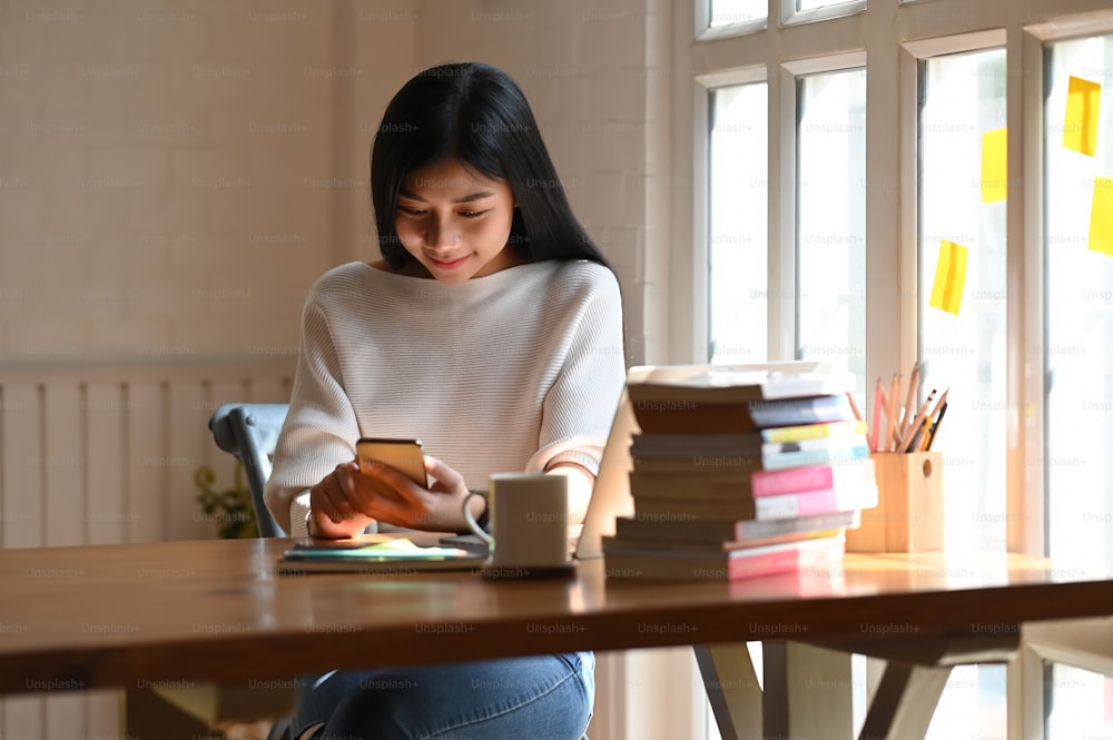 흰색 면 셔츠를 입은 젊은 아름다운 여성이 노트북, 커피 컵, 연필꽂이, 나무 테이블 위에 놓인 책 더미와 함께 앉아 스마트폰을 손에 들고 사용하고 있다.