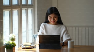 Frau benutzt / tippt auf einem Computer-Tablet, während sie vor einer Topfpflanze und Büchern am modernen Holztisch mit gemütlichem Wohnzimmer und Fenstern als Hintergrund sitzt.