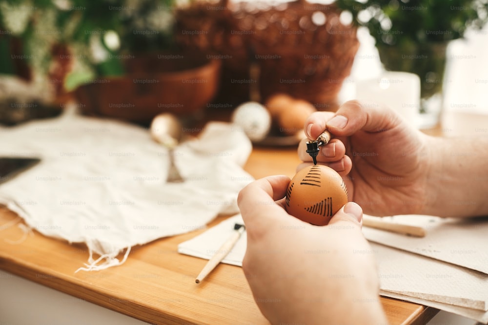 Painting Easter egg with hot wax on background of rustic wooden table with candle, basket, greenery. Easter egg with modern ornament in hand. Happy Easter. Ukrainian traditional pysanka.