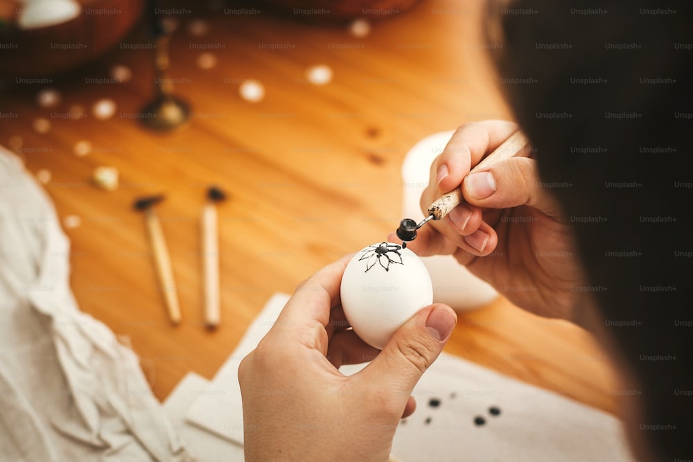 Mani che dipingono l'uovo di pasqua con cera calda sullo sfondo del tavolo di legno. Uova di Pasqua moderne con eleganti ornamenti di cera. Buona Pasqua. Zero sprechi. Pysanka tradizionale ucraina all'uovo