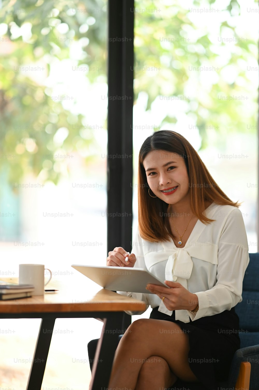 Portrait of young beautiful woman working as secretary using a computer tablet with stylus pen while sitting at the wooden working table with orderly office wall glass as background.