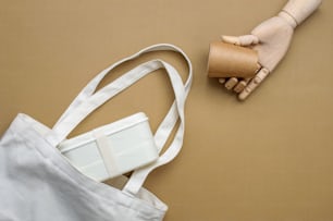 Wooden hand holding brown kraft paper cup near cotton bag with lunchbox on beige background. Top view, flat lay.