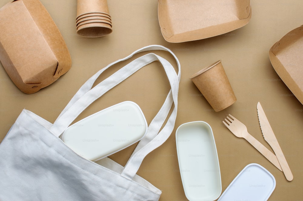 Disposable eco friendly food packaging. Brown kraft paper food containers, cups and lunchbox in fabric bag on beige background. Top view, flat lay.