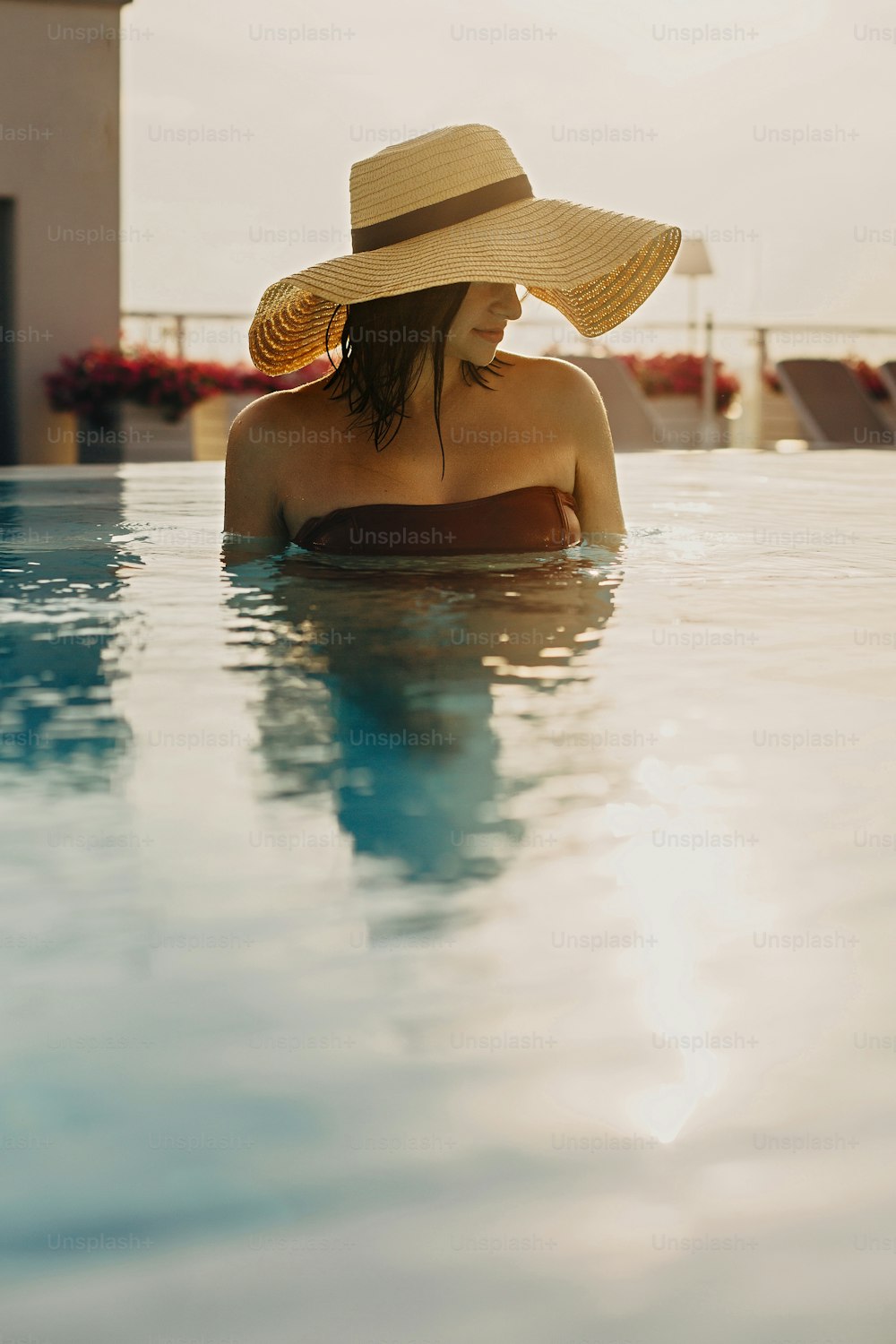 Stylish young woman in hat relaxing in pool and enjoying warm sunny light on summer vacation. Girl in sunhat on vacation in luxury tropical resort, swimming in pool on rooftop