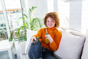 Young woman with curly red hair holding remote control and watching tv, changing channels. Cute girl drinking orange juice and watching tv in her apartment