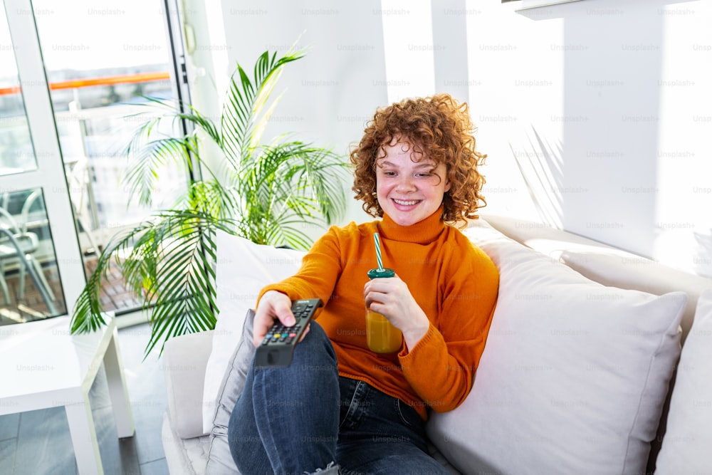 Young woman with curly red hair holding remote control and watching tv, changing channels. Cute girl drinking orange juice and watching tv in her apartment