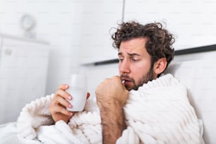 風邪と冬のインフルエンザウイルスに苦しむパジャマを着てベッドに横たわっている病気の無駄な男は、ヘルスケアのコンセプトで薬の錠剤を持っていますコップ一杯の水で丸薬を飲む