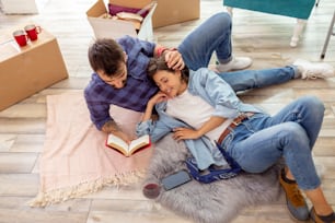 Couple in love moving in their new home, taking a break and relaxing, lying on the floor, drinking wine and reading a book