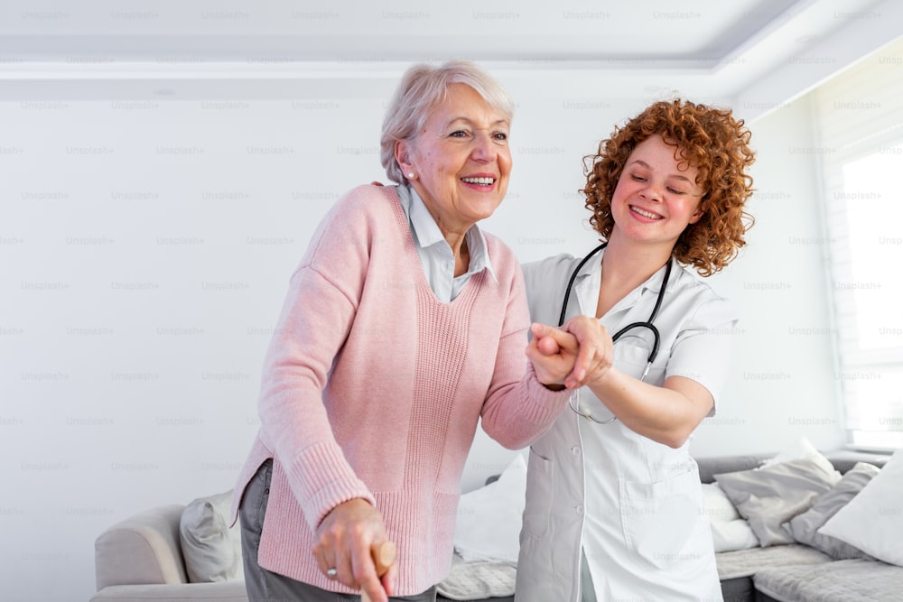 Enfermeira sorridente ajudando a senhora idosa a andar ao redor da casa de repouso. Retrato da cuidadora feliz e da mulher idosa caminhando juntos em casa. Profissional cuidador cuidando de idosa.