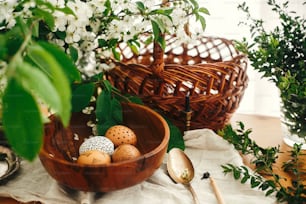 Natural Easter eggs painted with wax in wooden bowl on background of wicker basket, white spring flowers and greenery on wooden table. Happy Easter. Rural still life. Eco holiday. Zero waste