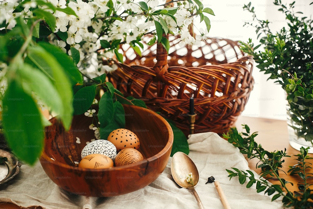 Huevos de Pascua naturales pintados con cera en cuenco de madera sobre fondo de cesta de mimbre, flores blancas de primavera y vegetación sobre mesa de madera. Felices Pascuas. Bodegón rural. Vacaciones ecológicas. Residuo cero
