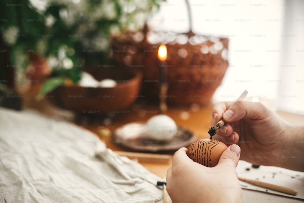 Painting Easter egg with hot wax on background of rustic wooden table with candle, basket, greenery. Easter egg with modern ornament in hand. Happy Easter. Ukrainian traditional pysanka. Zero waste