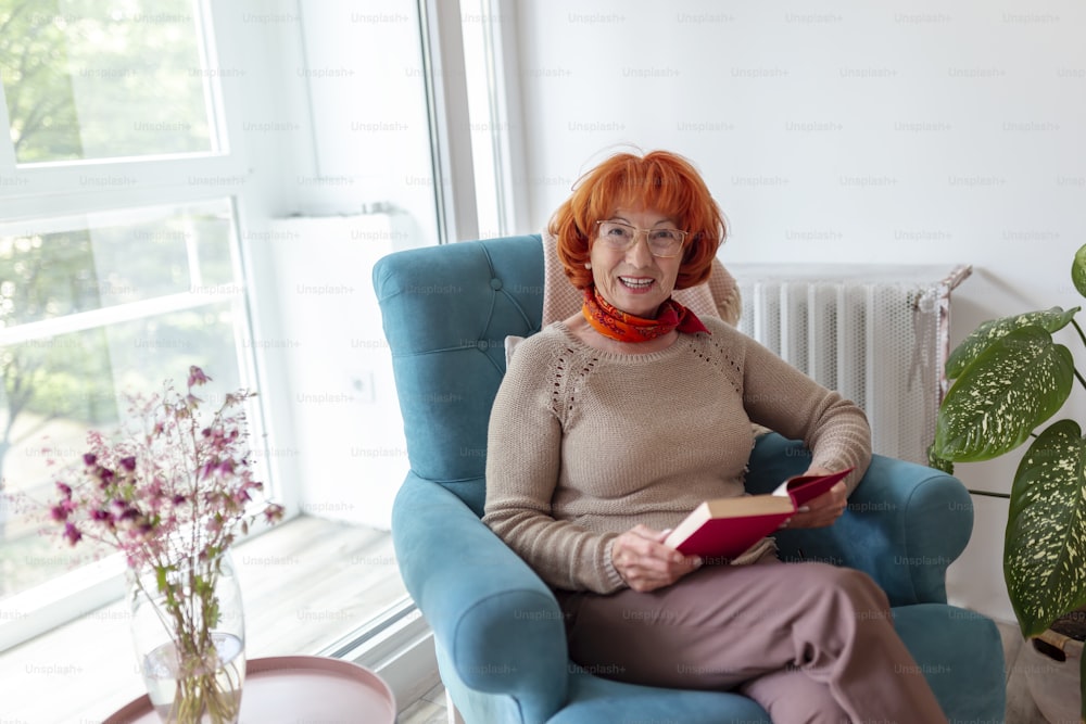 Elderly woman sitting in an armchair by the window, reading a book and relaxing at home