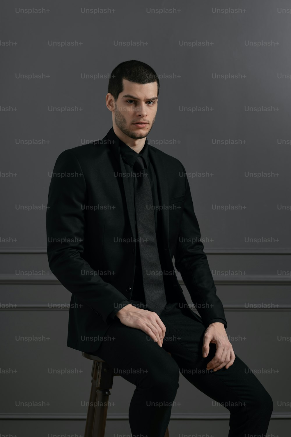 Junger gutaussehender Geschäftsmann in schwarzem Hemd und schwarzem Anzug. Selbstbewusster junger Geschäftsmann posiert auf grauem Hintergrund auf Stuhl sitzend